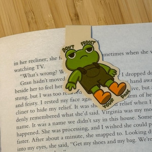 Pode incluir: Um marcador de livros caprichoso com um desenho de sapo de desenho animado e o texto "DON'T FROGET YOUR PAGE". O sapo é verde, com macacão marrom e botas laranja. O marcador de livros está em um livro aberto.