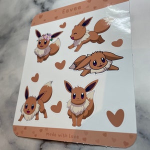 K&ouml;nnte beinhalten: Ein Blatt mit braunen und wei&szlig;en Aufklebern mit f&uuml;nf verschiedenen Illustrationen von Eevee, einem Pok&eacute;mon-Charakter. Die Aufkleber sind von kleinen Herzen und dem Text "Eevee by lex" und "made with love" umgeben.