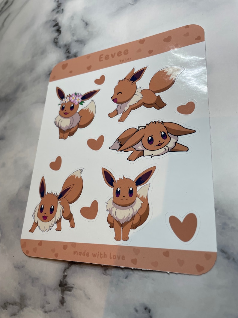 K&ouml;nnte beinhalten: Ein Blatt mit braunen und wei&szlig;en Aufklebern mit f&uuml;nf verschiedenen Illustrationen von Eevee, einem Pok&eacute;mon-Charakter. Die Aufkleber sind von kleinen Herzen und dem Text "Eevee by lex" und "made with love" umgeben.