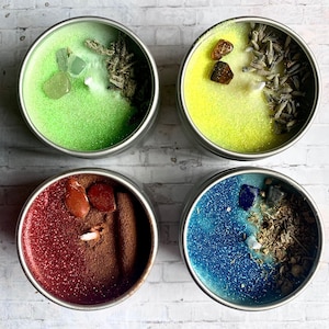 May include: Four small, round candles in silver tins. Each candle has a different color: green, yellow, red, and blue. The candles are decorated with glitter, crystals, and dried herbs. The candles are on a white brick background.