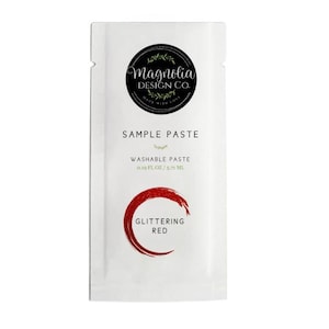 May include: A white packet of Magnolia Design Co. sample paste. The packet features a black circular logo with text and a red glitter paste sample. The text on the packet reads "SAMPLE PASTE", "WASHABLE PASTE", and "GLITTERING RED".