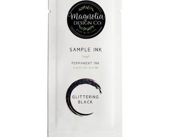 May include: A white sample ink packet from Magnolia Design Co. with the text "SAMPLE INK" and "GLITTERING BLACK". The packet also includes the text "PERMANENT INK 0.19 FL. OZ / 5.71 ML" and a black circular design.