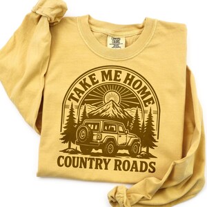 May include: Mustard-colored long-sleeve shirt with a brown graphic. The graphic features a mountain scene with a jeep and the text "TAKE ME HOME COUNTRY ROADS" in a circular design. The shirt is folded.