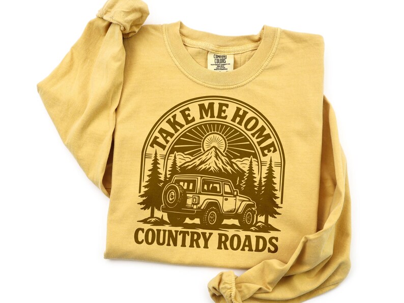 May include: Mustard-colored long-sleeve shirt with a brown graphic. The graphic features a mountain scene with a jeep and the text "TAKE ME HOME COUNTRY ROADS" in a circular design. The shirt is folded.