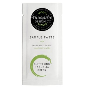May include: A white packet of Magnolia Design Co. sample paste. The packet has a black circular logo at the top with the words "Magnolia Design Co." and "Made with Love." Below the logo is the text "Sample Paste." The bottom of the packet has the words "Glittering Magnolia Green."