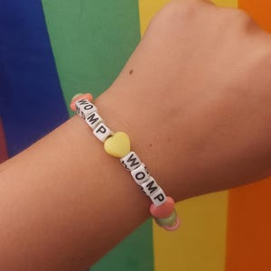 Puede incluir: Una pulsera de cuentas con la palabra "WOMP" escrita con cuentas de letras blancas. La pulsera incluye cuentas amarillas en forma de corazón y cuentas de acento rosas y verdes. El fondo es una bandera arcoíris.