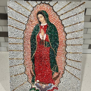 May include: A square artwork depicting Our Lady of Guadalupe, covered in sparkling crystals. The Virgin Mary is shown with hands clasped in prayer, wearing a red dress and green cloak. The image is surrounded by a halo of light.