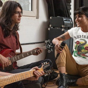 May include: Three people are in a room, two are playing guitars and one is holding a microphone. The person holding the microphone is wearing a white t-shirt with the word "AUSTRALIA" and a crocodile graphic.