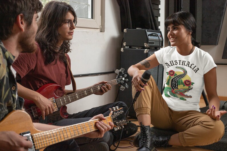 May include: Three people are in a room, two are playing guitars and one is holding a microphone. The person holding the microphone is wearing a white t-shirt with the word "AUSTRALIA" and a crocodile graphic.