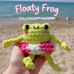 May include: A crocheted frog toy with a pink and white floatation device. The frog is light green with black eyes and a smiling mouth. The text "Floaty Frog PDF Crochet Pattern" is above the frog. The frog is held in front of a beach background.