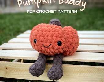 Pode incluir: Um brinquedo de amigo de abóbora de crochê com um rosto sorridente e pernas marrons. O brinquedo está sentado em uma caixa de madeira com grama verde no fundo. O texto "Pumpkin Buddy PDF CROCHET PATTERN" é exibido acima do brinquedo.