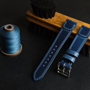 以下が含まれることがあります： ステッチのディテールが施された濃い青色の革製の腕時計用ストラップが2本。1本のストラップにはバックルが付いており、もう1本のストラップはストレートエンドになっています。画像には青い糸の糸巻きも写っています。