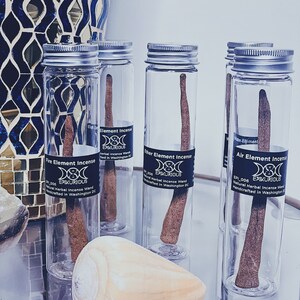 May include: Clear glass tubes with silver lids, each containing a brown incense wand. The tubes are labeled with "Fire Element Incense", "Ether Element Incense", and "Air Element Incense". A seashell rests on the glass surface.
