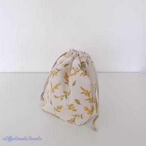 May include: A beige drawstring bag featuring a floral design of yellow flowers and green leaves. The bag has a neutral-coloured drawstring closure. The bag is made of fabric and is set against a white background.