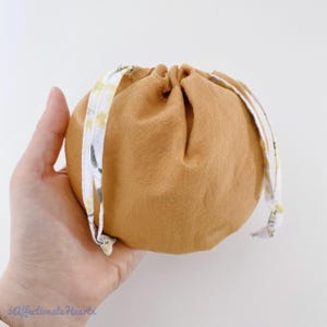 以下が含まれることがあります： 温かみのあるマスタードイエローの、小さくて丸い巾着袋。 手で持っており、白地に黄色とグレーの繊細な花柄の紐で開閉します。