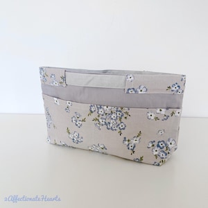 May include: A rectangular fabric organiser with a floral pattern. The light grey fabric features blue and white flowers. The organiser has a grey band and two pockets. The top has two grey handles. The text "2 Affectionate Hearts" is at the bottom.
