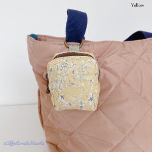 May include: A small, yellow fabric pouch with a white floral pattern and a brown zip, attached to a dusty pink quilted tote bag with dark blue straps. The pouch features a metal ring connector and a small dark charm on the zip pull.