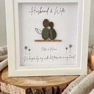 May include: A white framed artwork with the words "Husband & Wife" at the top. The artwork features a couple made of stones, with the names "Peter & Marie" below. The text "No longer by my side, but forever in my heart" is at the bottom.