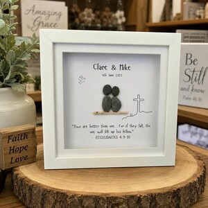 May include: A white framed artwork with the names "Clare & Mike" and the date "6th June 2015." The artwork features pebble figures, a cross, and a quote from Ecclesiastes. The frame is displayed on a wooden slice.