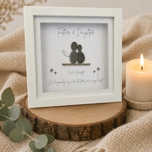 May include: A white framed pebble art piece with the words "Father & Daughter". The artwork depicts a father and daughter made of pebbles, with the names "Dad & Charlotte" and a quote below. The frame is displayed on a wooden slice.