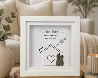 May include: A white framed artwork with the text "First Home", a house illustration, and the names "Martin & Melissa" with the date "30th April 2026". The artwork includes pebble art figures and star accents.