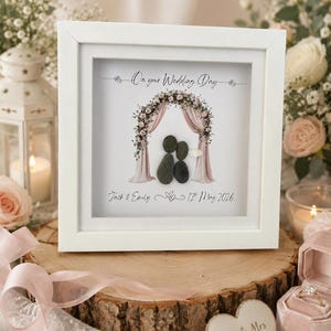 May include: White framed wedding art with pebble art couple under a floral arch. Text reads "On your Wedding Day", names "Jack & Emily" and a date. The artwork has soft pink and white floral accents. A romantic keepsake.