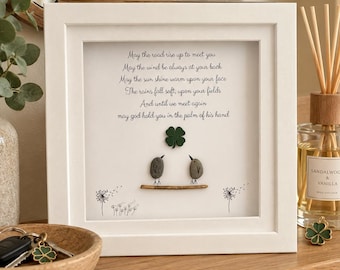 May include: A square white frame containing pebble art of two gray birds perched on a twig beneath a green four-leaf clover. The text reads: May the road rise up to meet you, May the wind be always at your back, May the sun shine warm upon your face, The rains fall soft upon your fields, And until we meet again, may god hold you in the palm of his hand.