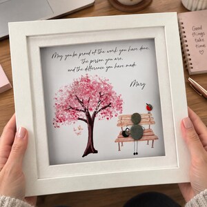 May include: A square white frame containing a pebble art piece featuring a painted pink cherry blossom tree, a bench, and a figure made of grey stones. The text reads: May you be proud of the work you have done, the person you are, and the difference you have made. Mary.