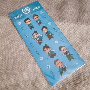 Pu&ograve; includere: Un foglio di adesivi azzurro con illustrazioni di samurai in stile chibi. Gli adesivi presentano varie pose ed espressioni. Sul foglio sono visibili testo giapponese e un emblema circolare.