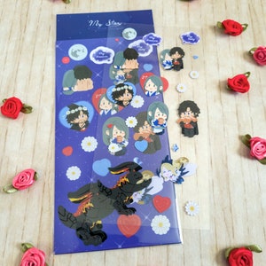 Pu&ograve; includere: Un foglio di adesivi con sfondo blu e la scritta "My Star". Gli adesivi raffigurano coppie di cartoni animati, cuori, lune e un drago nero. Piccoli adesivi a forma di margherita bianca e accenti di rose rosse sono sparsi.