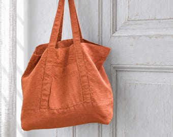 May include: A large, burnt orange tote bag featuring two long shoulder straps and a horizontal seam detail across the lower section. The bag has a visible exterior pocket on the front panel and is constructed with a textured fabric finish.