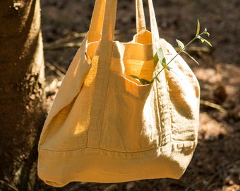 May include: A large, light yellow canvas tote bag with two long handles. The bag is hanging from a tree branch and has a small green plant sticking out of the top.