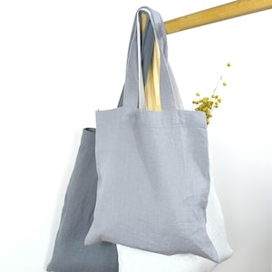 Könnte beinhalten: Zwei Tragetaschen aus Leinenstoff. Die Tasche links ist grau und die Tasche rechts ist weiß. Beide Taschen haben lange Henkel.