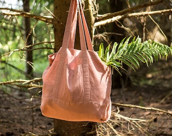 May include: A pink linen tote bag hanging from a tree branch in a forest setting. The bag has a large pocket on the front and is made of a soft, natural fabric.