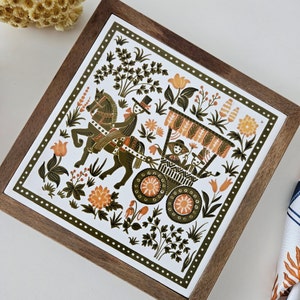 Puede incluir: Un azulejo de cerámica blanco con marco de madera presenta una ilustración colorida de un carruaje tirado por un caballo con personas dentro. El diseño incluye motivos florales y una combinación de colores verde, marrón y blanco.