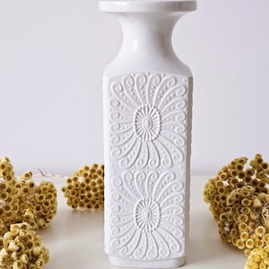 Op de afbeelding: Witte keramische vaas met een vierkant lichaam en een brede, uitlopende opening. De vaas is versierd met een reliëfpatroon van gestileerde bloemen.