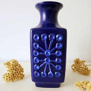 Może przedstawiać: Kobaltowy, ceramiczny wazon o prostokątnym korpusie i rozszerzającej się szyi. Wazon posiada wypukły, stylizowany kwiatowy wzór w jaśniejszym odcieniu niebieskiego. Wazon ma około 25 cm wysokości.