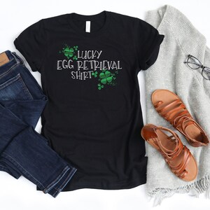 Puede incluir: Camiseta negra con el texto "Lucky Egg Retrieval Shirt" en blanco con diseños de trébol verde.