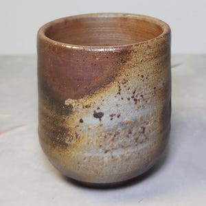 Peut inclure: Une tasse en céramique faite à la main avec une esthétique rustique. La tasse présente un mélange de teintes marron, beige et blanche, avec une surface texturée. Le design est simple et cylindrique, adapté aux boissons ou à la décoration.