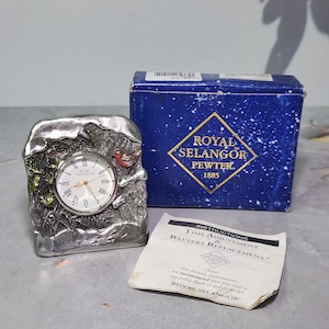 Puede incluir: Un reloj de escritorio de peltre con una esfera redonda y números romanos. El reloj está enmarcado en un marco decorativo y texturizado con un pájaro y follaje. Una caja azul con las palabras "Royal Selangor Pewter" está detrás del reloj.