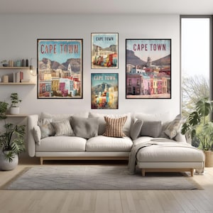 Peut inclure: Quatre impressions d'art encadrées avec le texte "CAPE TOWN" en rouge et blanc, représentant des bâtiments colorés et des paysages de montagne. Les impressions sont affichées au-dessus d'un canapé blanc avec des coussins gris, créant un cadre de salon confortable.