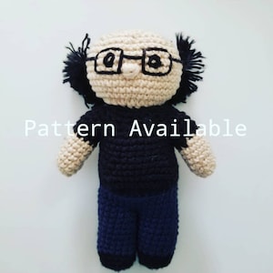 May include: Crocheted doll with black hair, glasses, and a black shirt and blue pants. The doll is wearing a black shirt and blue pants. The text "Pattern Available" is visible in the image.