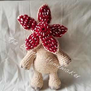 May include: A handmade crochet Demogorgon plush toy. The Demogorgon has a cream-colored body, arms, and legs. The head is a vibrant red with white accents. The text "The Ever Homemaker" is written on the left side of the image.