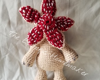 May include: A handmade crochet Demogorgon plush toy. The Demogorgon has a cream-colored body, arms, and legs. The head is a vibrant red with white accents. The text "The Ever Homemaker" is written on the left side of the image.