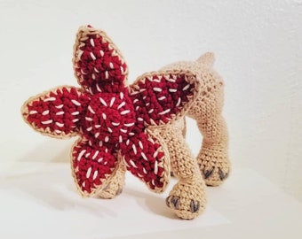 May include: A crocheted Demogorgon plush toy. The creature features a large, open, flower-like head in deep red with white accents. The body and legs are tan, with gray claws. The toy is handmade and sits on a white surface.