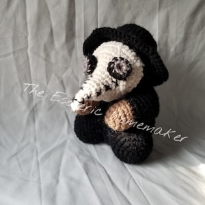 May include: A crocheted plush toy of a plague doctor wearing a black hat and a long white beak mask. The toy is sitting on a white fabric surface.