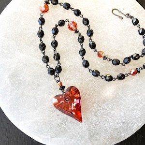 May include: A black beaded necklace with a red crystal heart pendant. The necklace features black faceted beads and small red crystal accents.