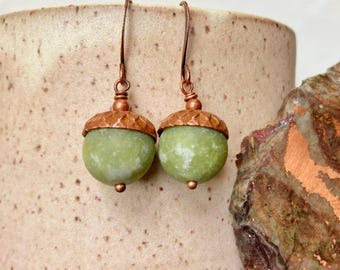 May include: A pair of dangle earrings featuring round, light green stone beads shaped like acorns. Each earring has a textured copper-colored metal cap and a matching copper-colored hook wire for suspension, creating a cohesive, nature-inspired design.