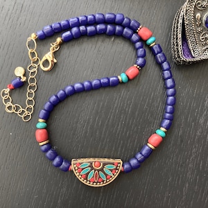Peut inclure: Un collier artisanal avec des perles bleu foncé, des perles couleur corail et des accents turquoise. Le collier a un fermoir doré et un pendentif décoratif avec des détails rouges, turquoise et dorés. Bijou pour un look élégant.