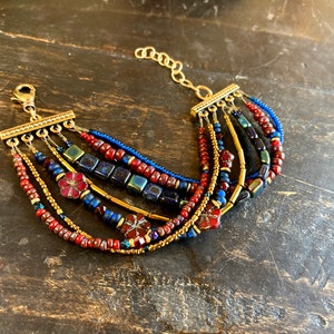 May include: Multi-strand bracelet with red, blue, and gold beads. The bracelet features a clasp closure and decorative flower-shaped beads.